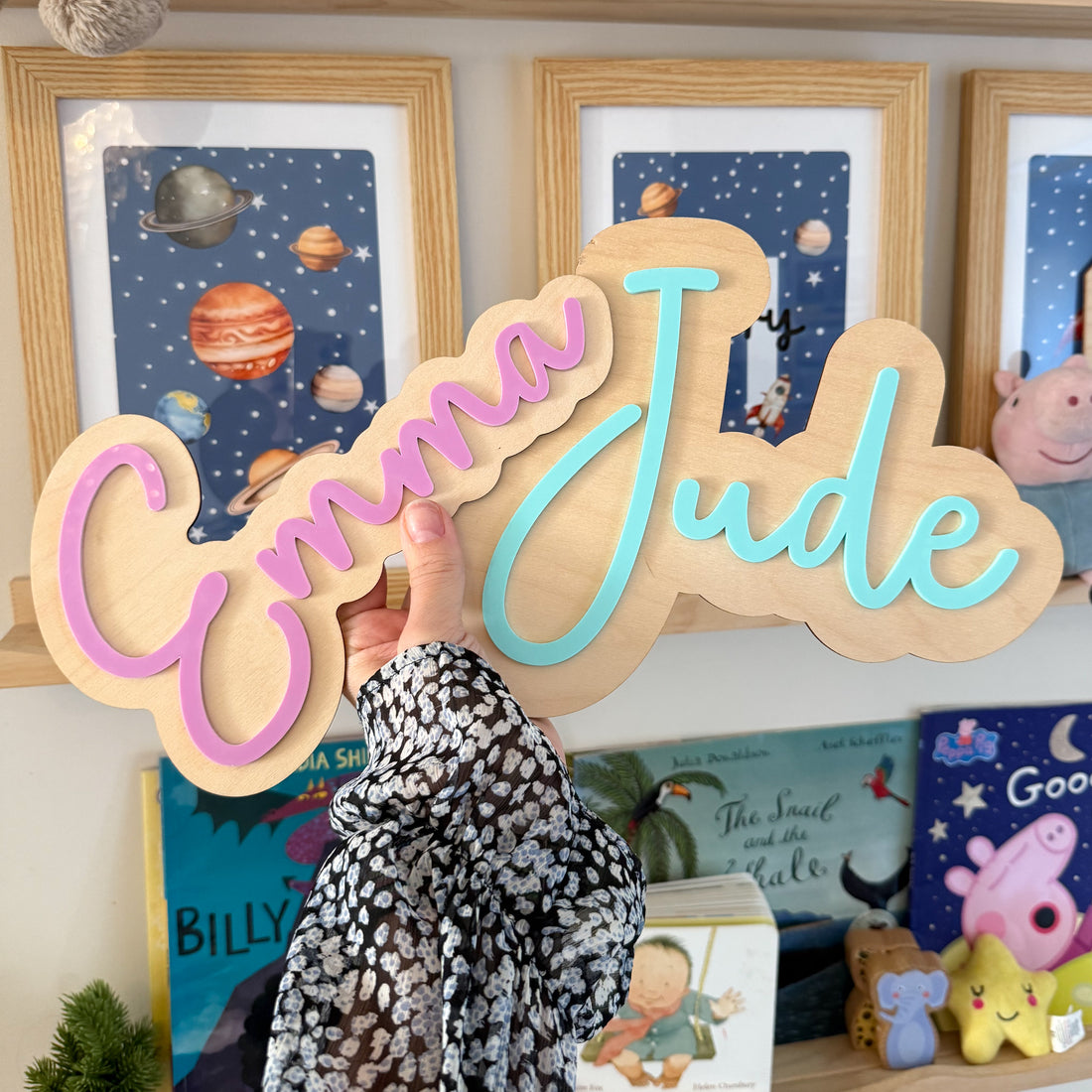 A person is holding a wooden sign with the name &quot;Emma Jude&quot; written on it, in front of a shelf with children&