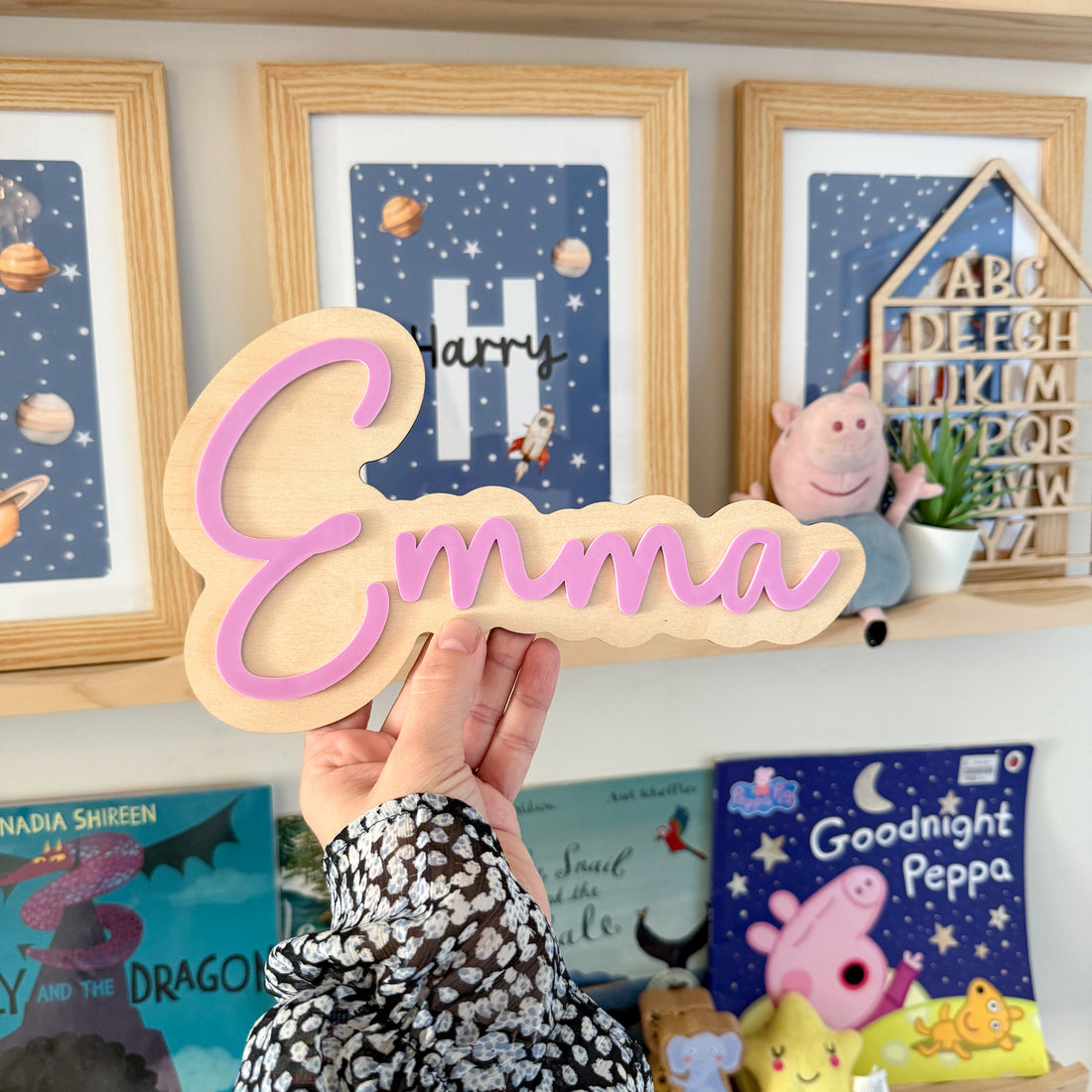 A person is holding a wooden sign with the name &quot;Emma&quot; written on it, in front of a shelf with children&